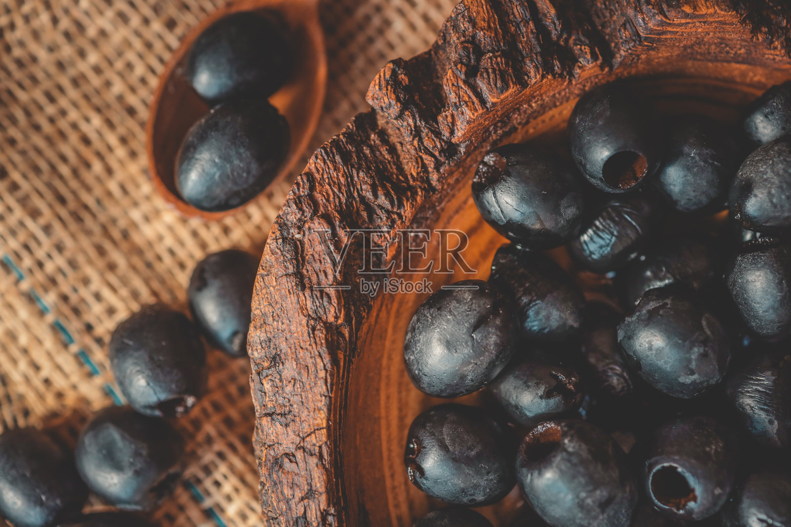 装满黑橄榄的乡村木碗和一把木勺，放在麻布上，沐浴在温暖的自然光中，适合地中海厨房或美食概念。照片摄影图片