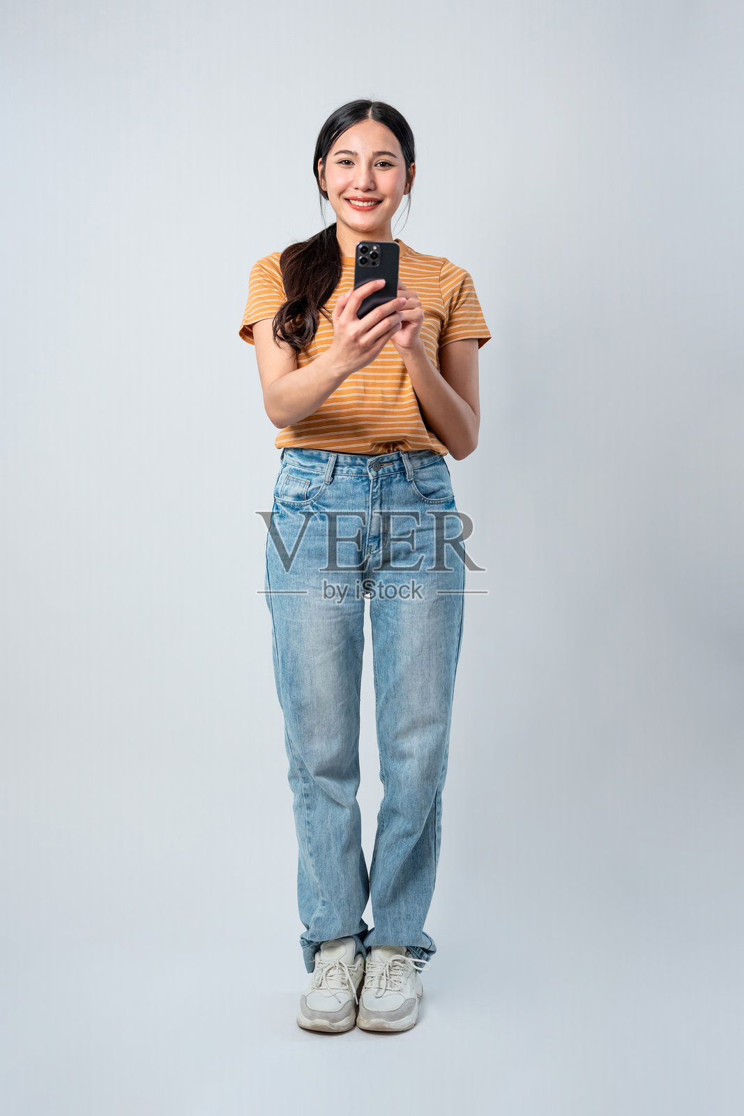 亚洲美丽女性在灰色背景下手持手机。迷人的年轻女孩模特在使用在线数字智能手机设备时感到快乐和愉悦。照片摄影图片