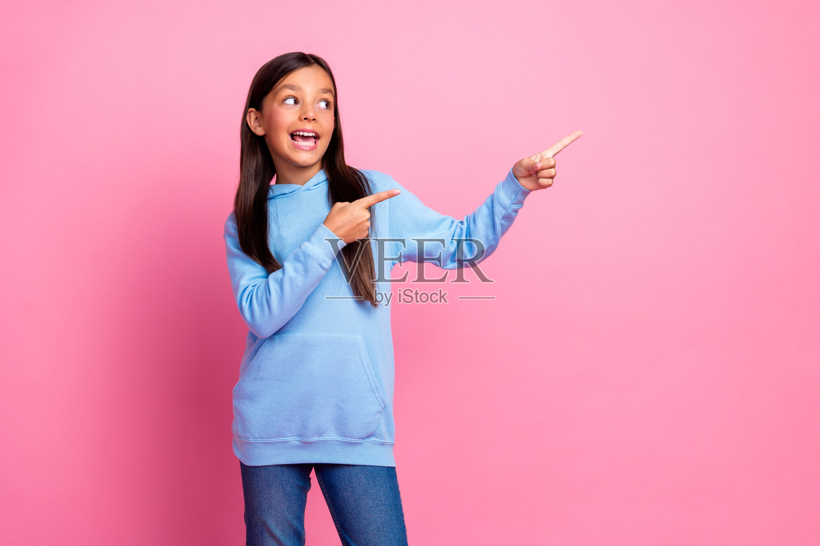 穿着蓝色连帽衫的快乐小女孩在鲜艳的粉色背景前兴奋地指着。照片摄影图片