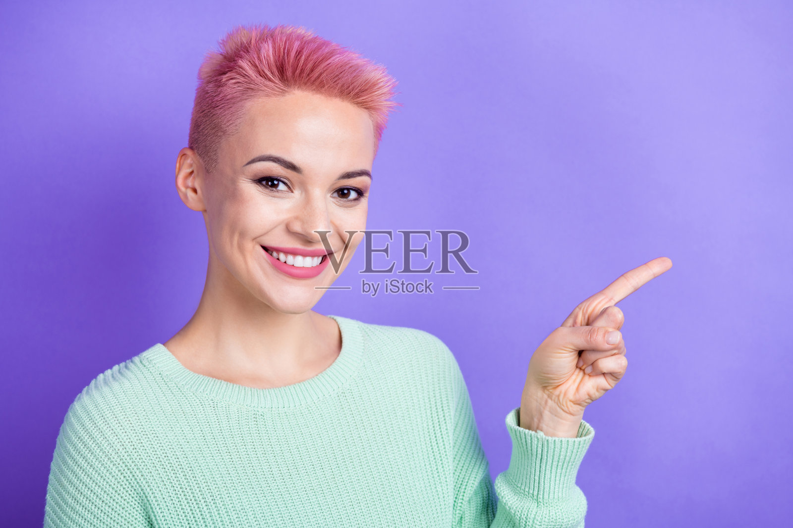 穿着青色毛衣的可爱积极女士，手指指向紫色背景中的空白处。照片摄影图片