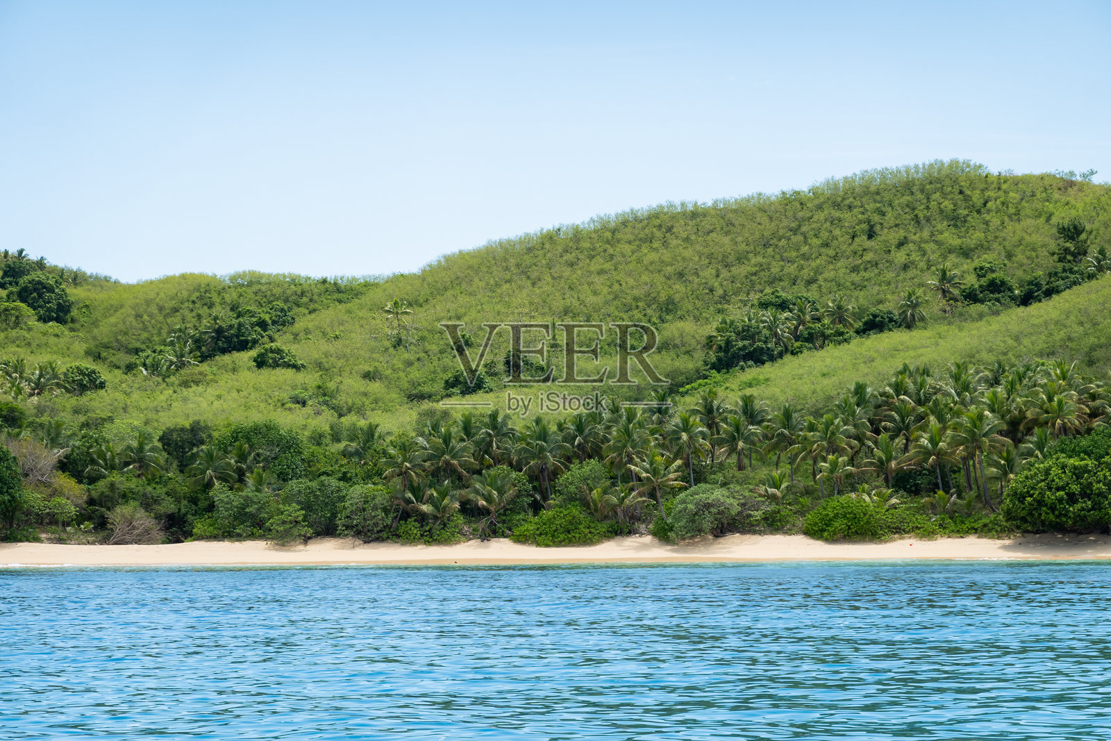 从船上看，碧绿色的海浪向热带岛屿延伸，生机勃勃的植被和棕榈树在灿烂的天空下沿海岸排成一排。照片摄影图片