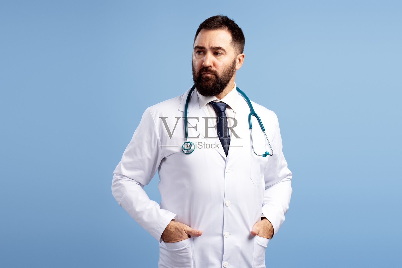 自信的医生侧身看着，双手插在口袋里，背景为蓝色。照片摄影图片