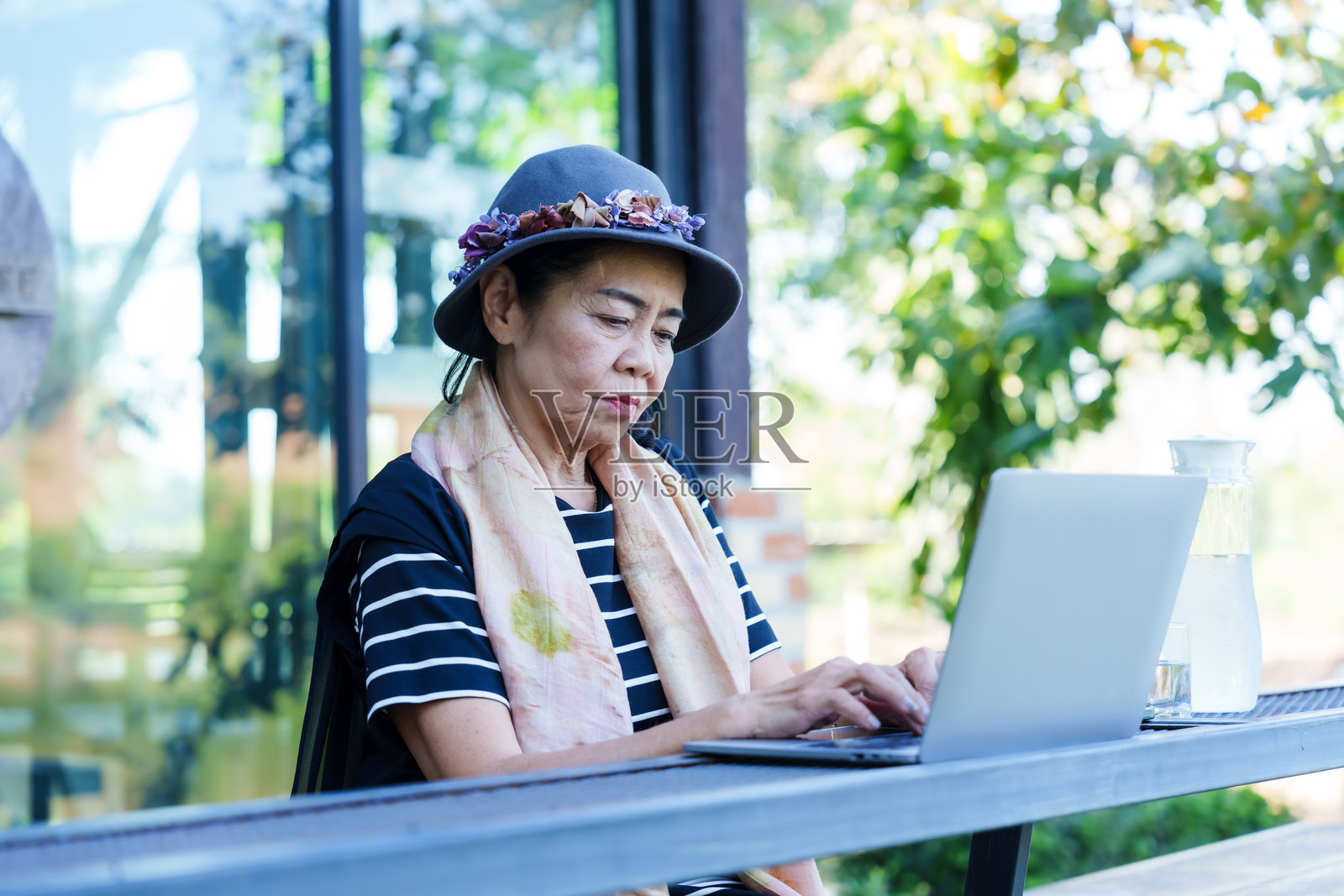 穿着休闲条纹衣服的亚洲老年女性在户外咖啡馆使用笔记本电脑，享受自然绿意，宁静的自由职业远程工作，享受独立、休闲和退休生活的快乐。照片摄影图片