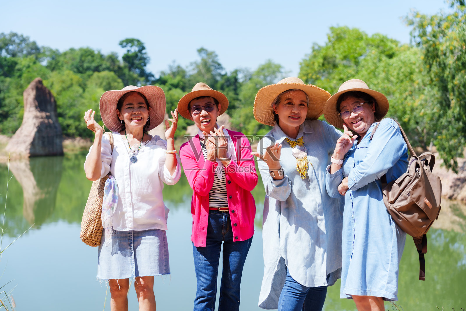 一群亚洲老年女性站在湖边，微笑着拍手，穿着色彩鲜艳的休闲服装，戴着太阳帽，背着背包，享受着阳光明媚的户外自然假期冒险。照片摄影图片