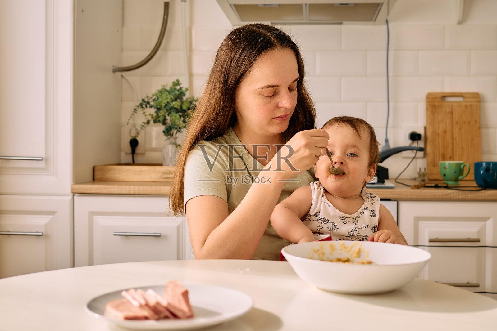 一位母亲在阳光明媚的厨房里温柔地喂养她的宝宝。孩子兴奋地张嘴准备吃一勺食物，突显了他们共同度过的温馨用餐时刻。照片摄影图片