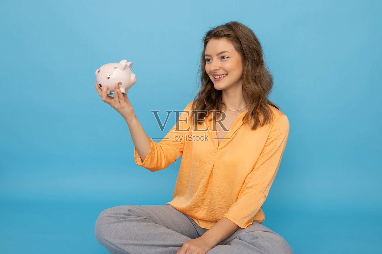 年轻女性盘腿坐着，手里拿着储蓄罐，微笑着看着自己的存款照片摄影图片