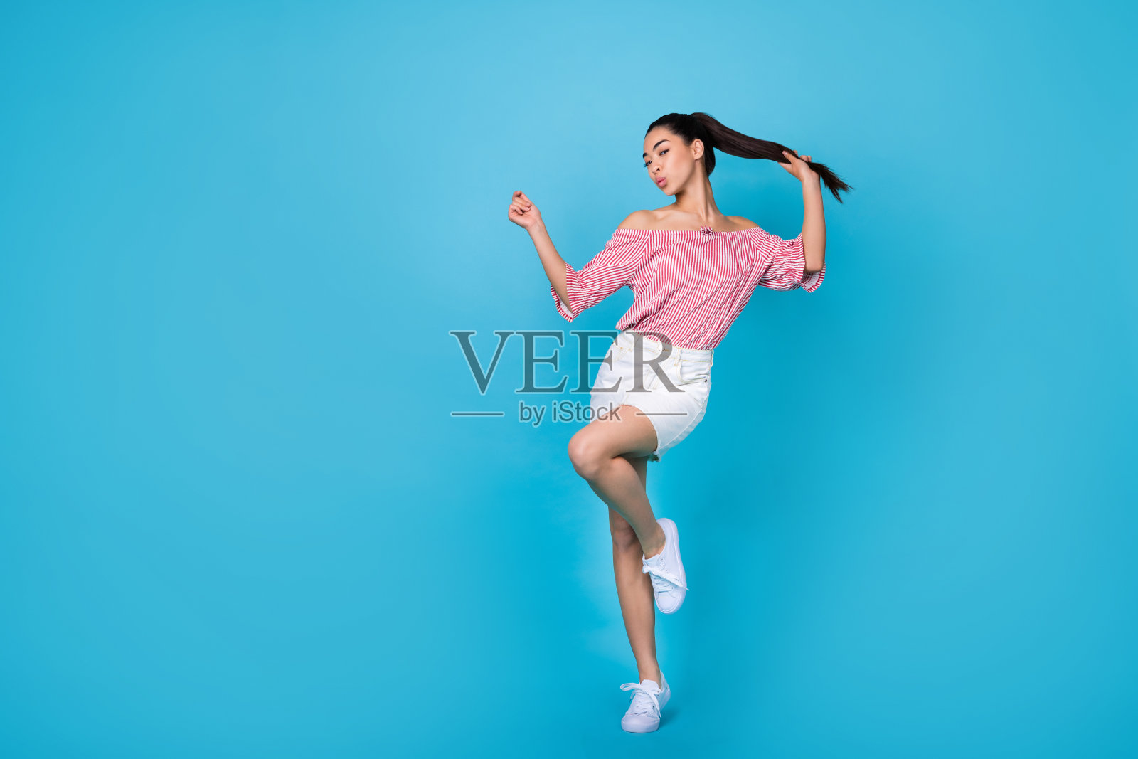 穿着露肩条纹衬衫的可爱时尚女孩在空旷的蓝色背景下跳舞，享受快乐时光。照片摄影图片