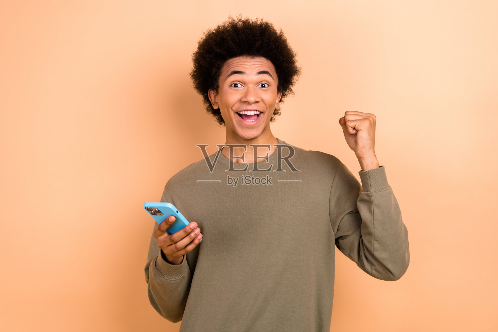 一位英俊快乐的男子手持智能手机，举起拳头庆祝成功，背景为米色。照片摄影图片