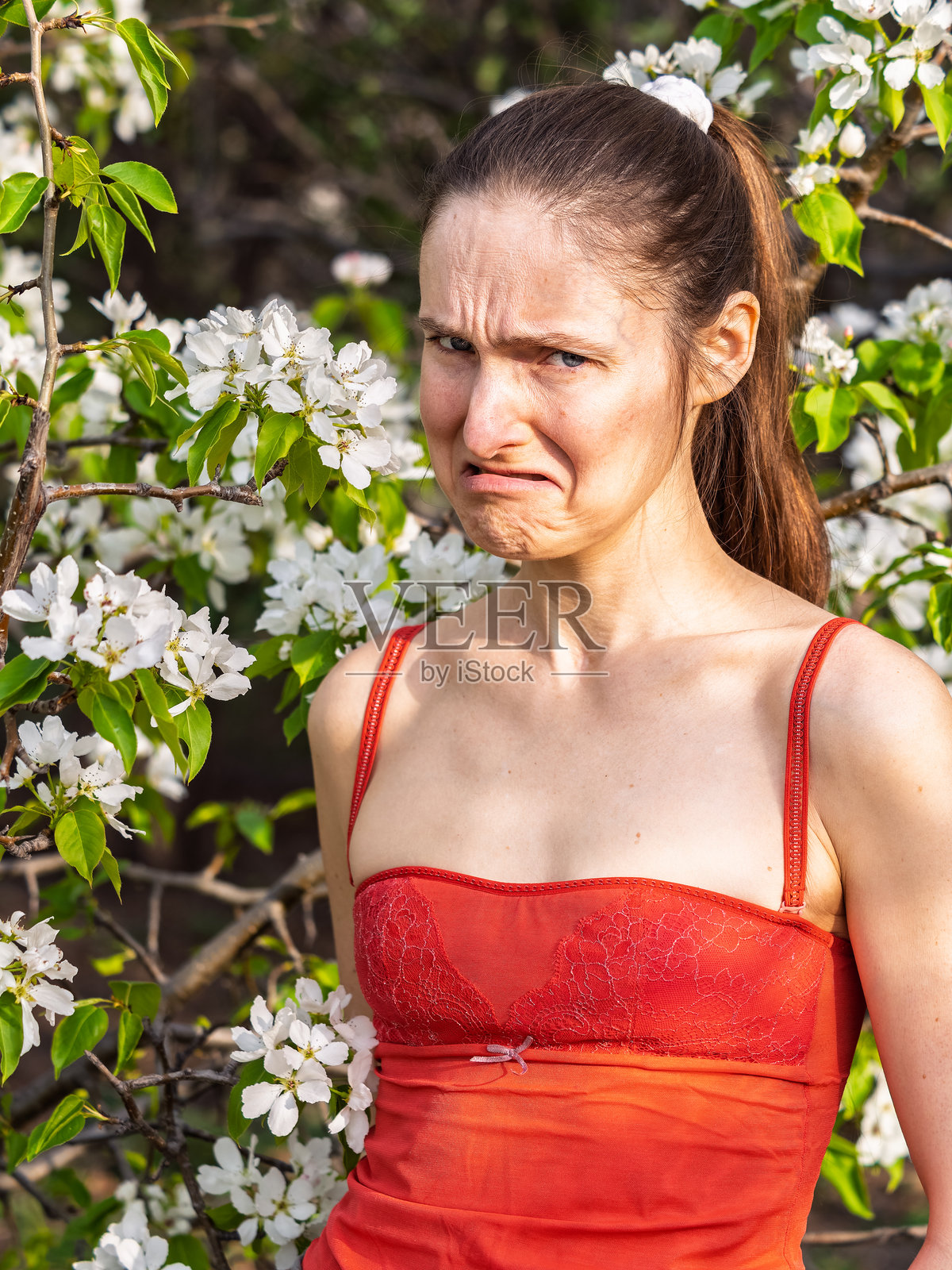 年轻女性在公园里打喷嚏，背景是一棵开花的树。花粉过敏概念。照片摄影图片