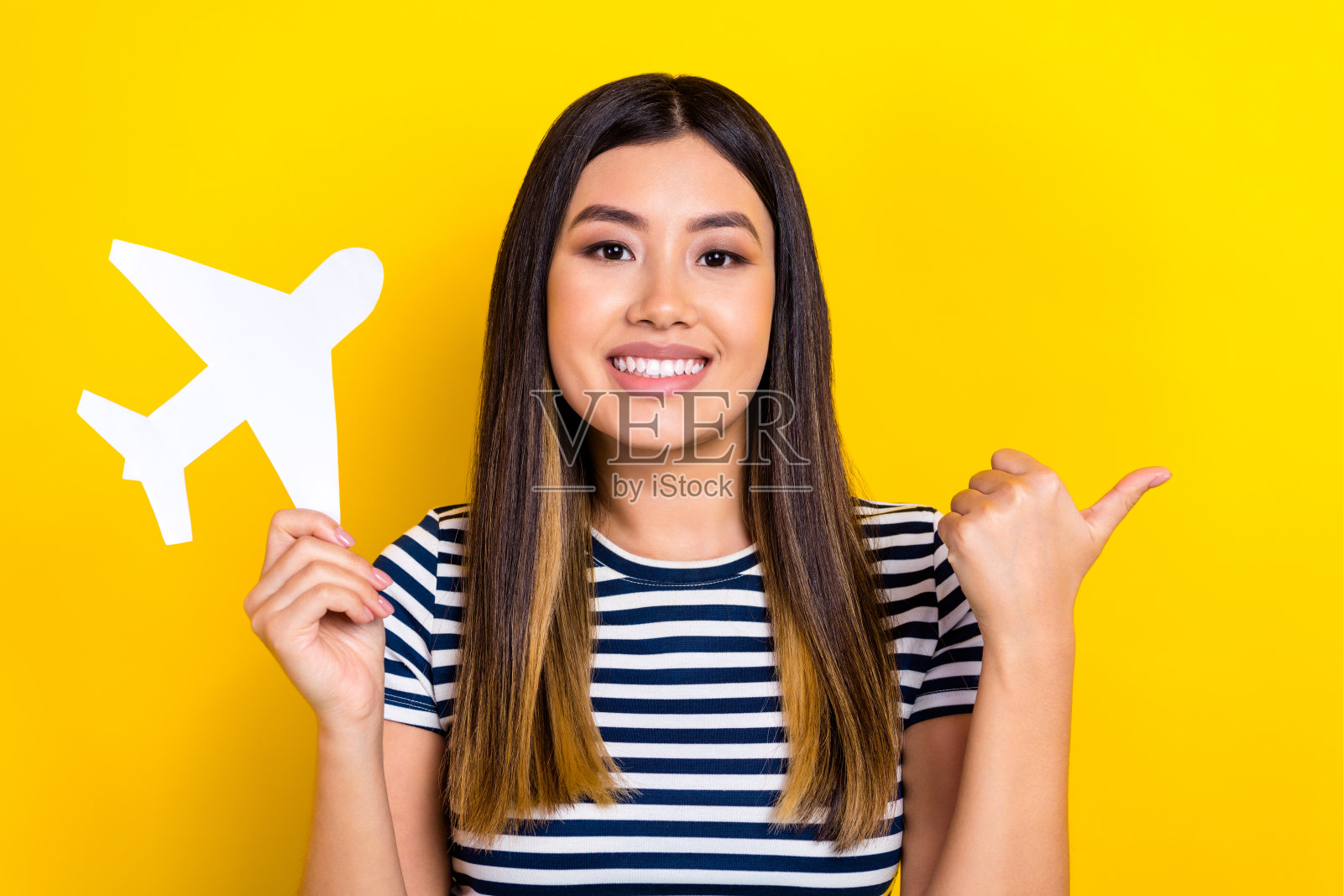 年轻的越南女士黑发，手持纸飞机，指向空旷的地方，背景为黄色，象征低成本航班。照片摄影图片