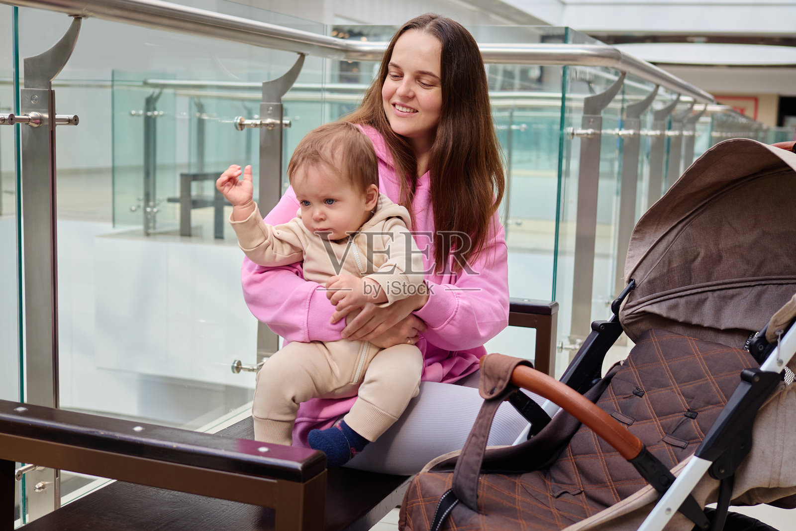 一位母亲坐在商场的长椅上，微笑着抱着她的小孩。这个幼儿穿着米色的衣服，好奇地四处张望，而婴儿车则静静地停在旁边。照片摄影图片