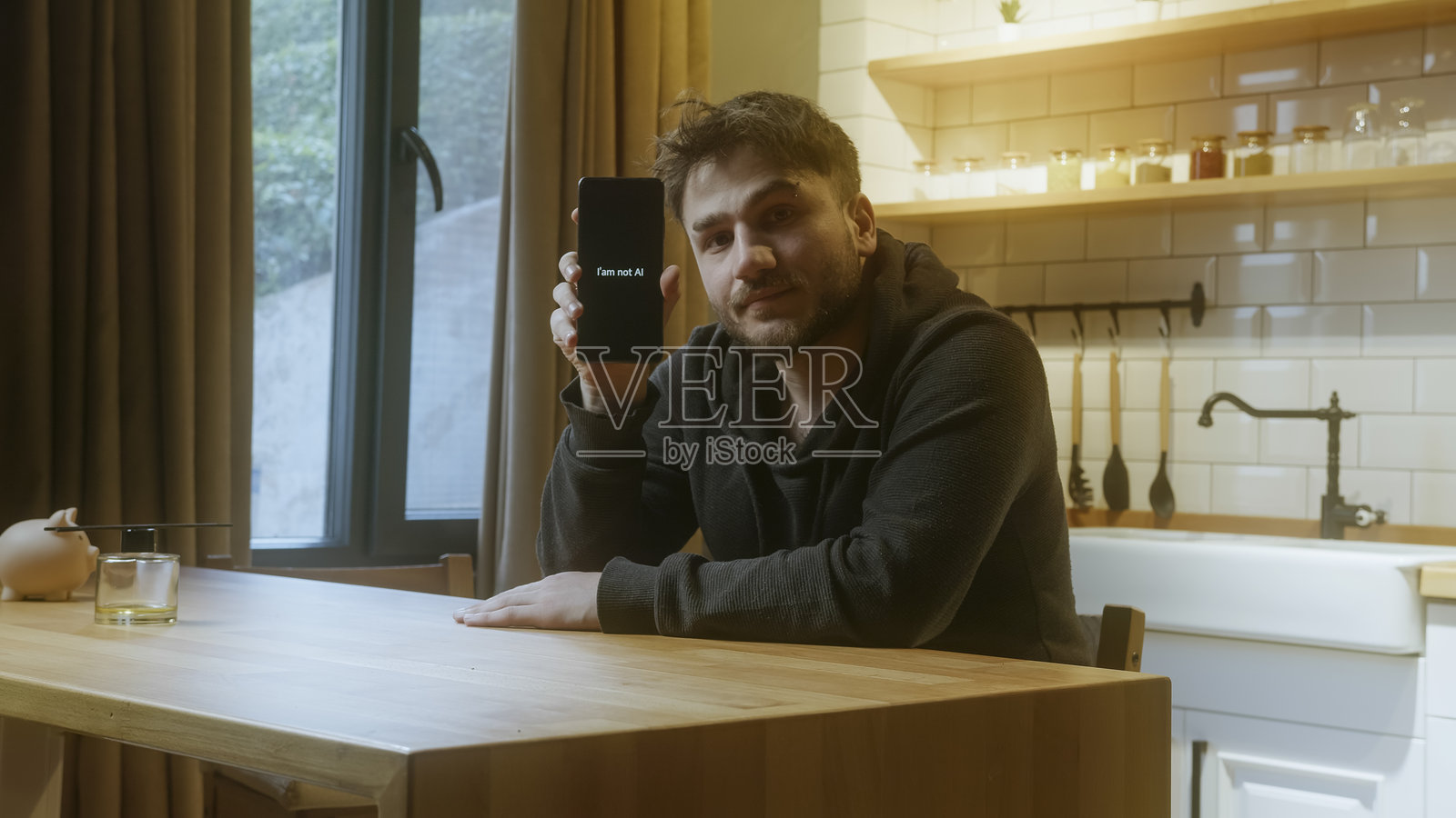 男人在舒适的厨房里手持手机，屏幕上显示“我不是人工智能”的信息，以强调人类的真实性。照片摄影图片