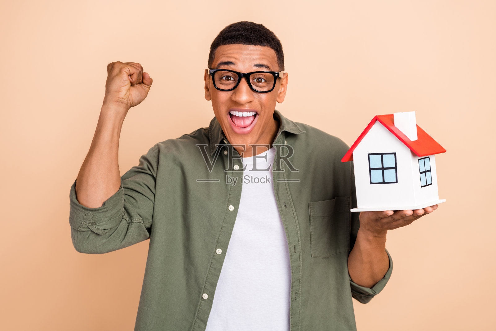 穿着卡其色衬衫、戴着眼镜的兴奋男士手持小房子庆祝搬家，背景为柔和色彩。照片摄影图片