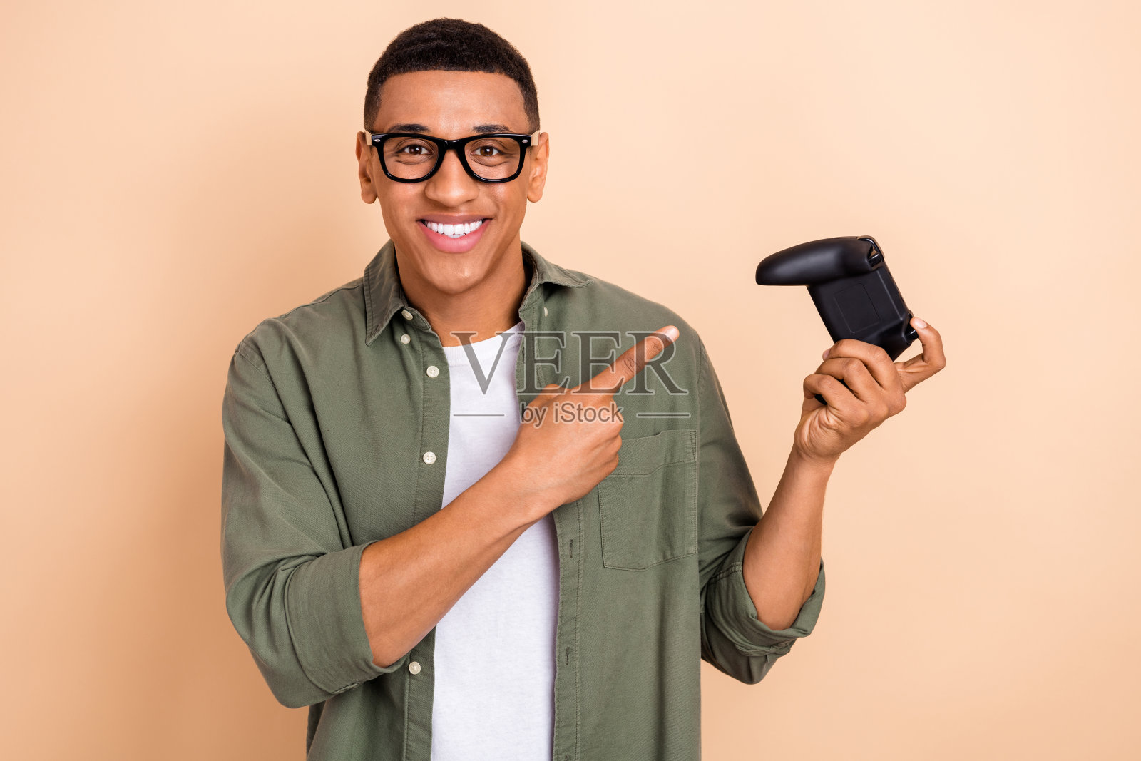 满意的男子肖像，穿着时尚衬衫和眼镜，指着游戏手柄，推荐购买游戏，背景为米色。照片摄影图片