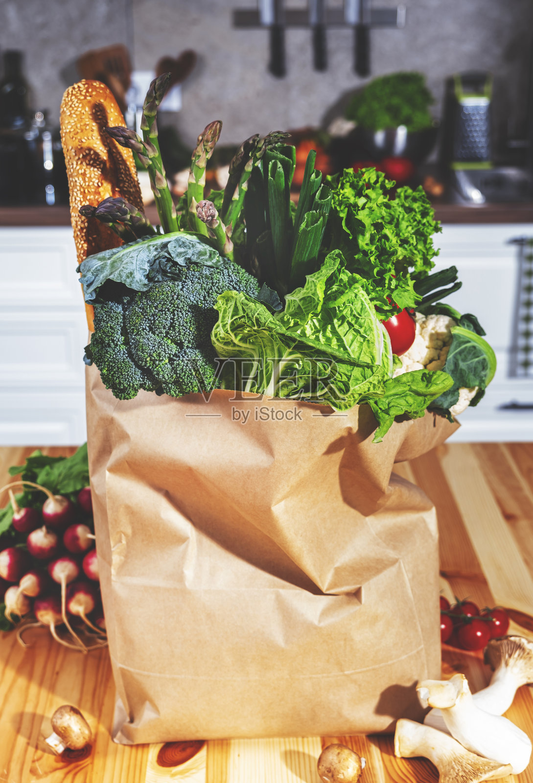 纸质环保袋装着新鲜食材，现代舒适厨房的木桌上摆放着健康午餐的食材，包括蔬菜、花椰菜、法棍、芦笋、西红柿和蘑菇。照片摄影图片