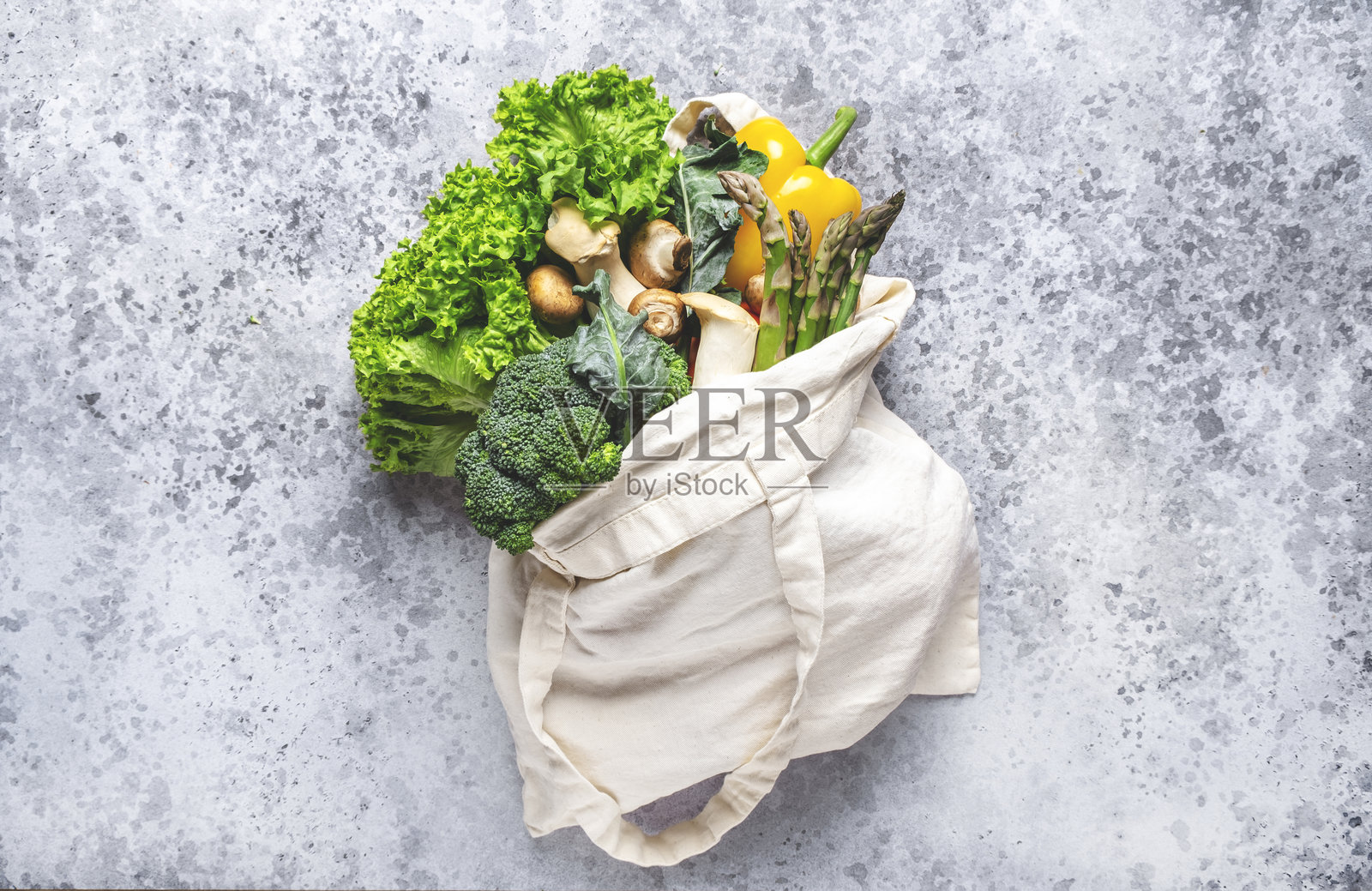 新鲜食品背景。生态购物袋里装着绿色蔬菜、香草、西兰花、芦笋和蘑菇，放在灰色桌子上，俯视图。照片摄影图片