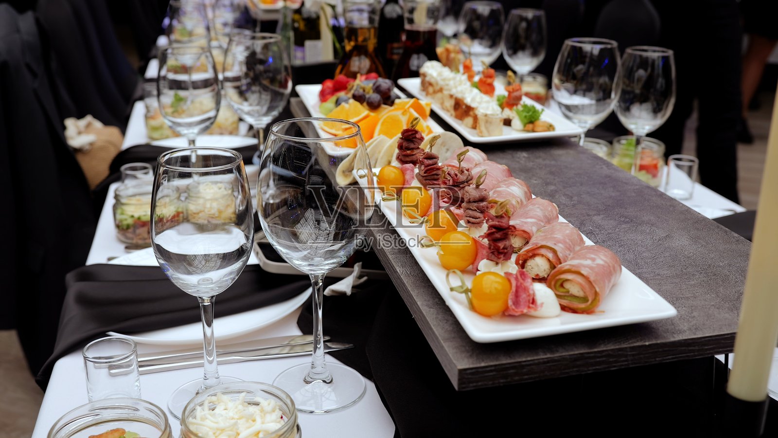 酒杯、开胃菜盘上摆放着小吃、肉卷和水果串，布置在黑白桌布上，突显优雅的宴会准备。照片摄影图片