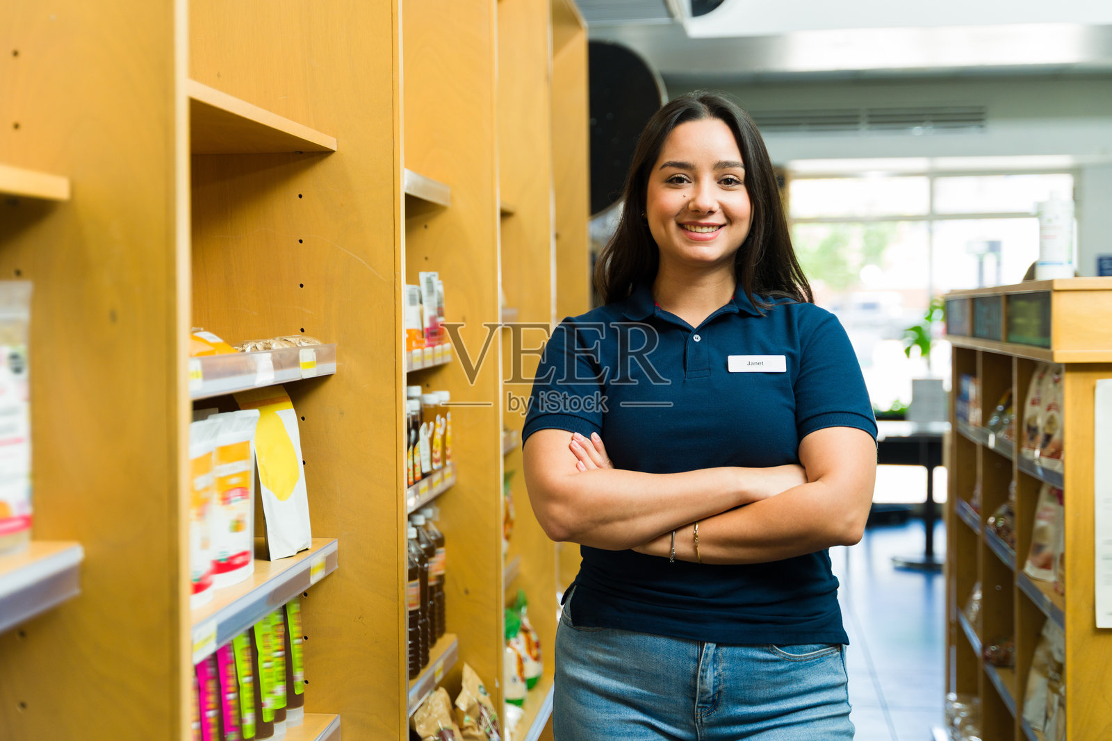 微笑的女销售员在超市过道中摆姿势，双臂交叉。照片摄影图片