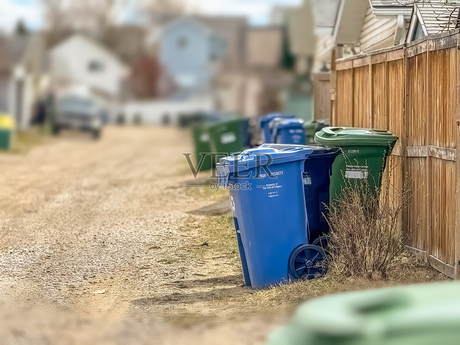 一个蓝色的回收箱矗立在一条土路上，旁边是一个木栅栏，背景中模糊可见其他垃圾箱和房屋。照片摄影图片
