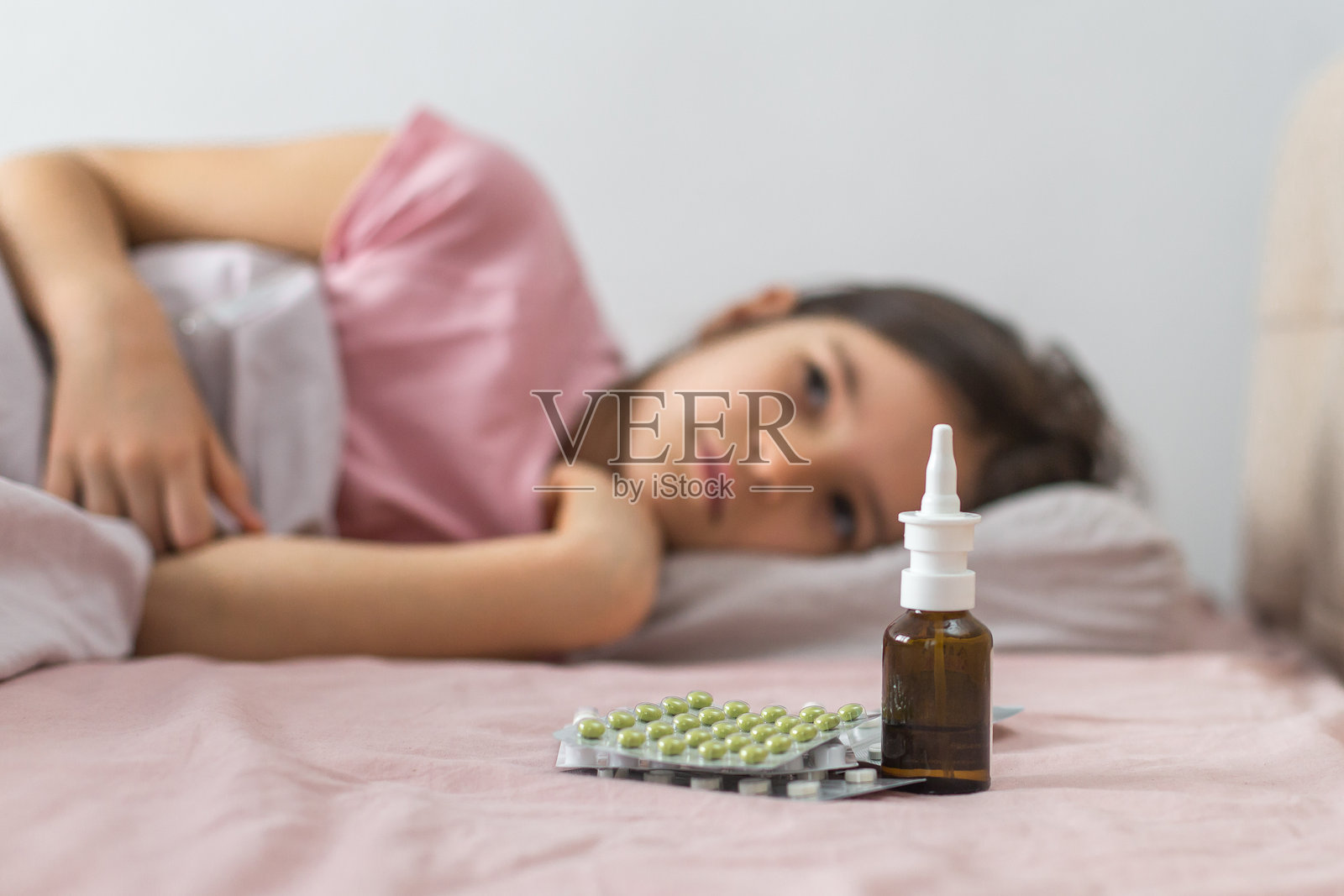 一位生病的女孩躺在床上，身边有药物、鼻用喷雾和绿色胶囊的泡罩包装，躺在粉色的毯子上，看起来疲惫且不适。疾病与康复的概念。照片摄影图片