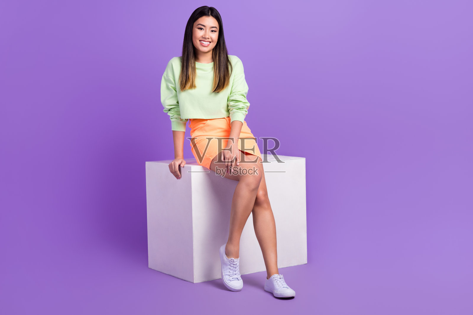 穿着绿色衬衫和时尚裙子的乐观友好的女性坐在白色立方体上，背景为紫色。照片摄影图片