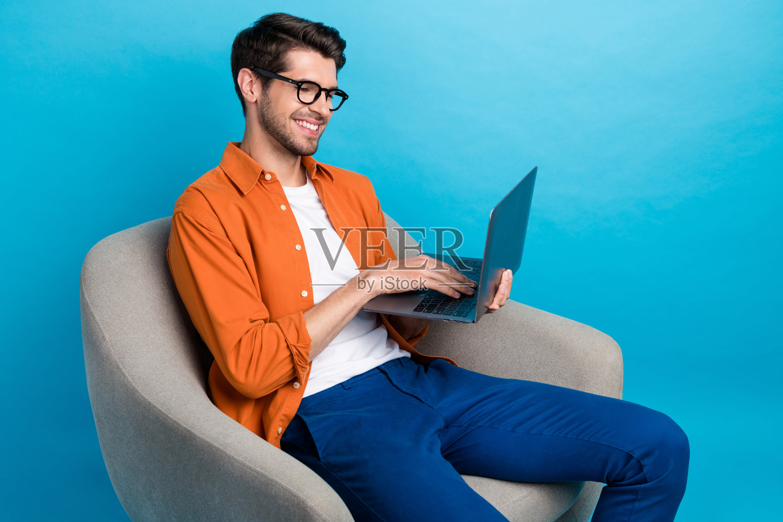 积极聪明的男士坐在舒适的椅子上，使用无线笔记本电脑进行远程工作，背景为蓝色。照片摄影图片