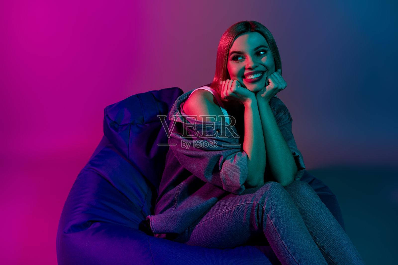 积极的千禧女孩坐在未来派咖啡馆的豆袋椅上，背景是生动的霓虹色光芒。照片摄影图片