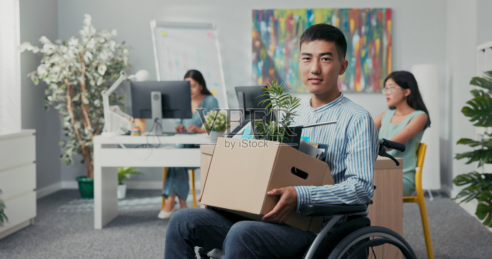 坐在轮椅上的男子手里拿着一盒工作必需品，开心地微笑着，背景中的同事们在庆祝。照片摄影图片