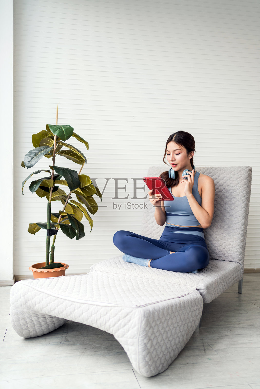 一位亚洲年轻女性盘腿坐在现代休闲椅上，戴着耳机阅读一本书，旁边有一盆植物，房间明亮且极简。戴着耳机的女性享受着她的平板电脑。照片摄影图片