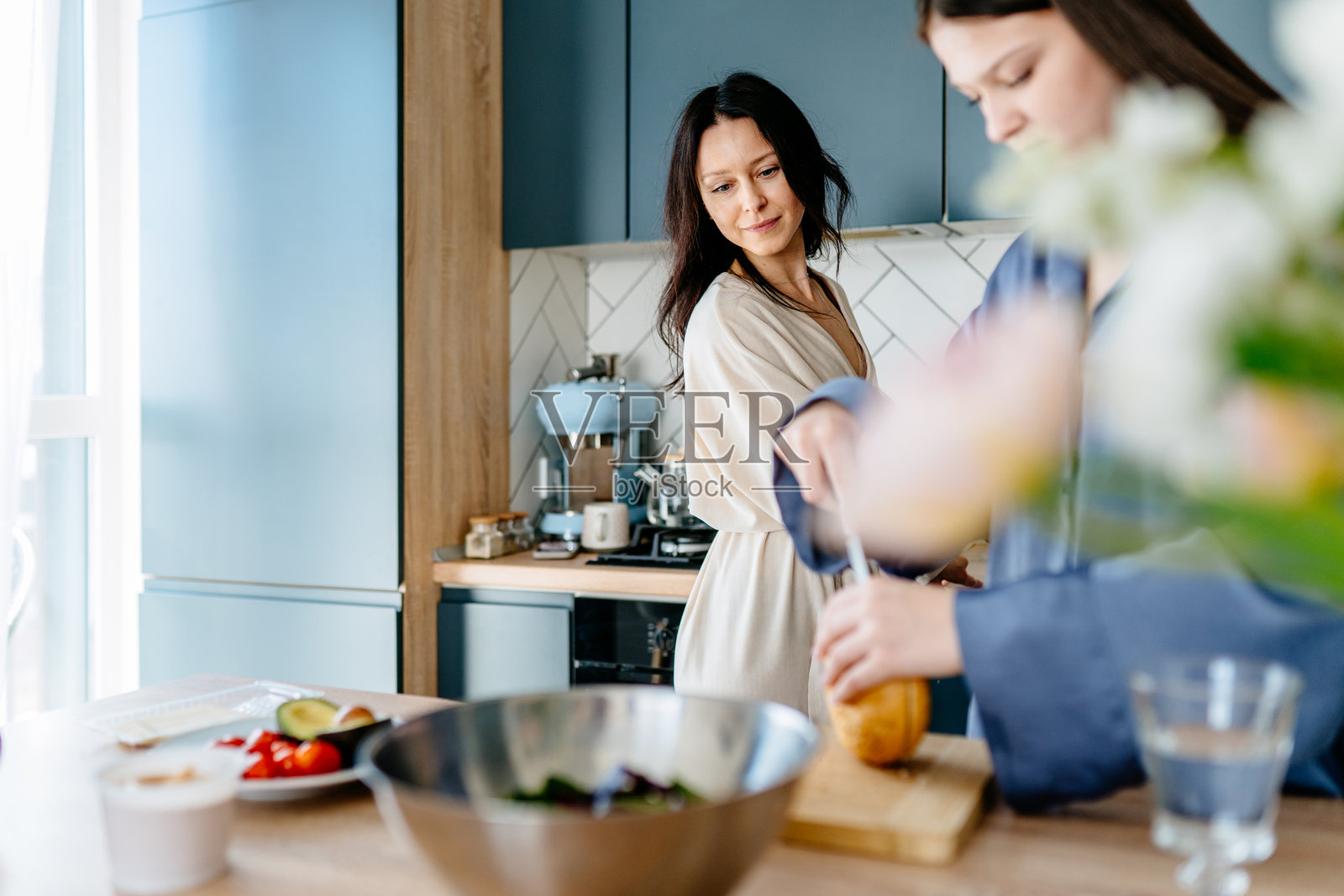 母亲在温馨的厨房里看着青少年女儿切面包，柔和的晨光洒在周围。照片摄影图片