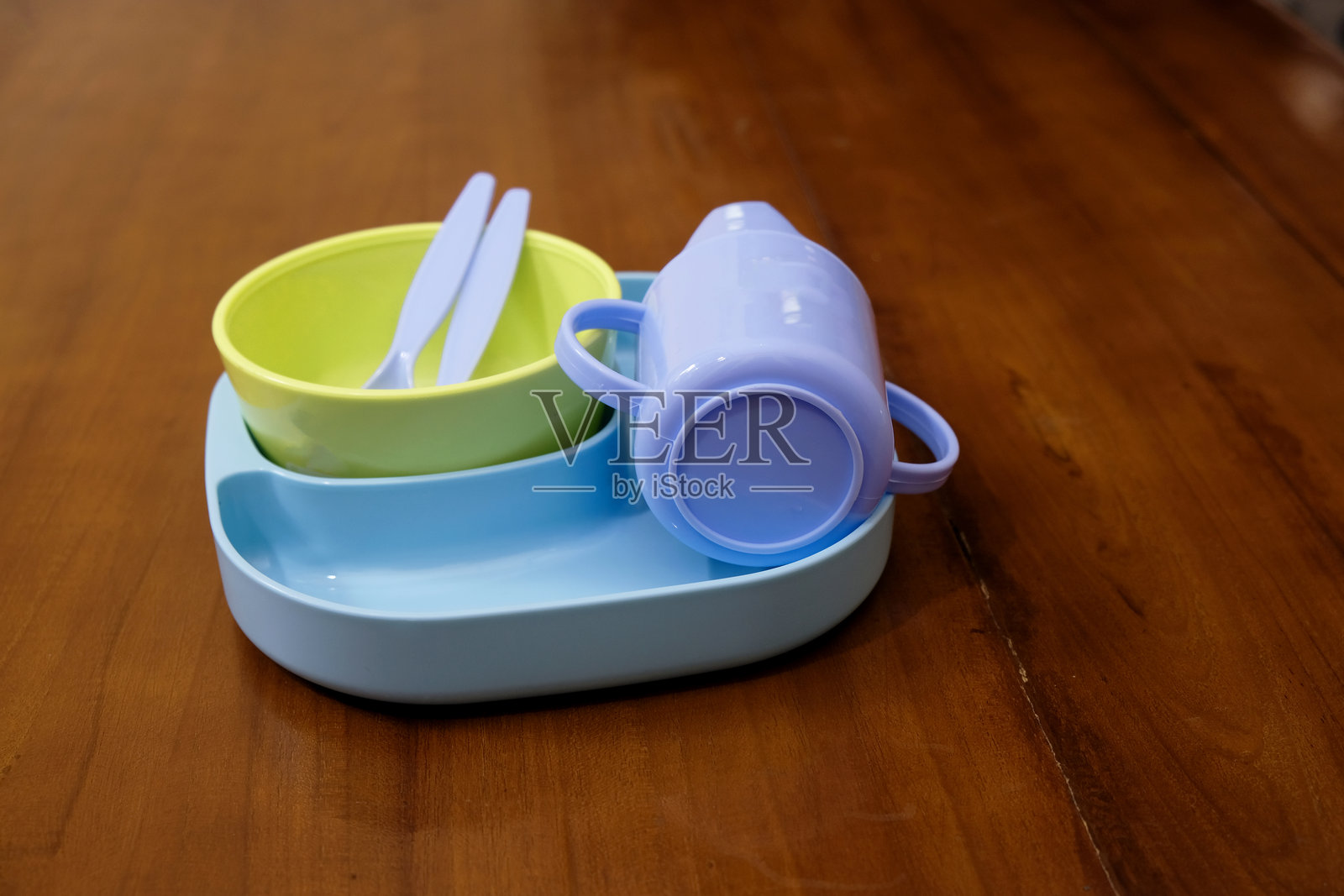 塑料盘子和碗，勺子和叉子，以及杯子。婴儿餐具放在木桌背景上。照片摄影图片