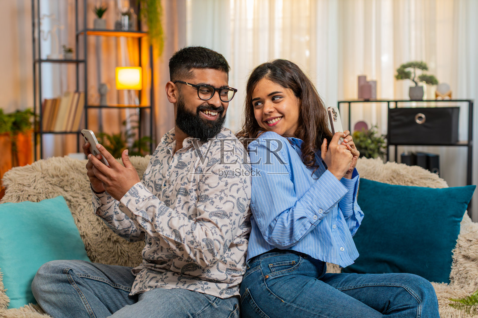 印度家庭夫妻，男女背靠背，手持智能手机，微笑着一起观看搞笑视频。照片摄影图片