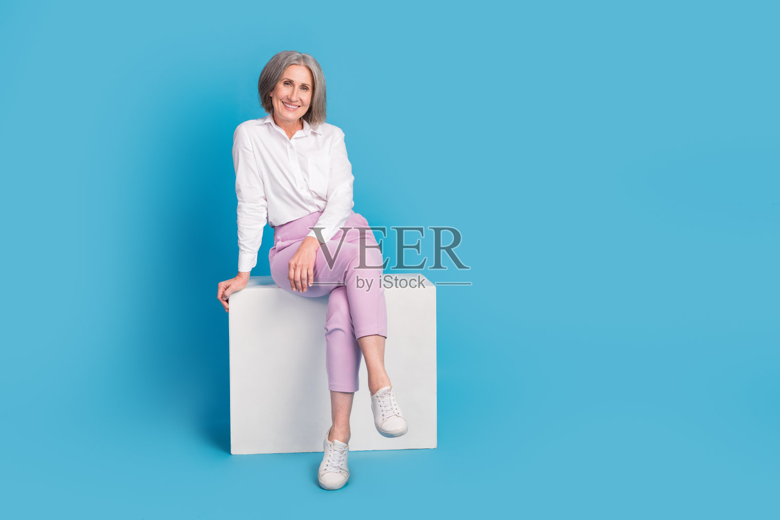 全身肖像，积极可爱的人坐在讲台上，摆姿势，背景为空旷的蓝色。照片摄影图片