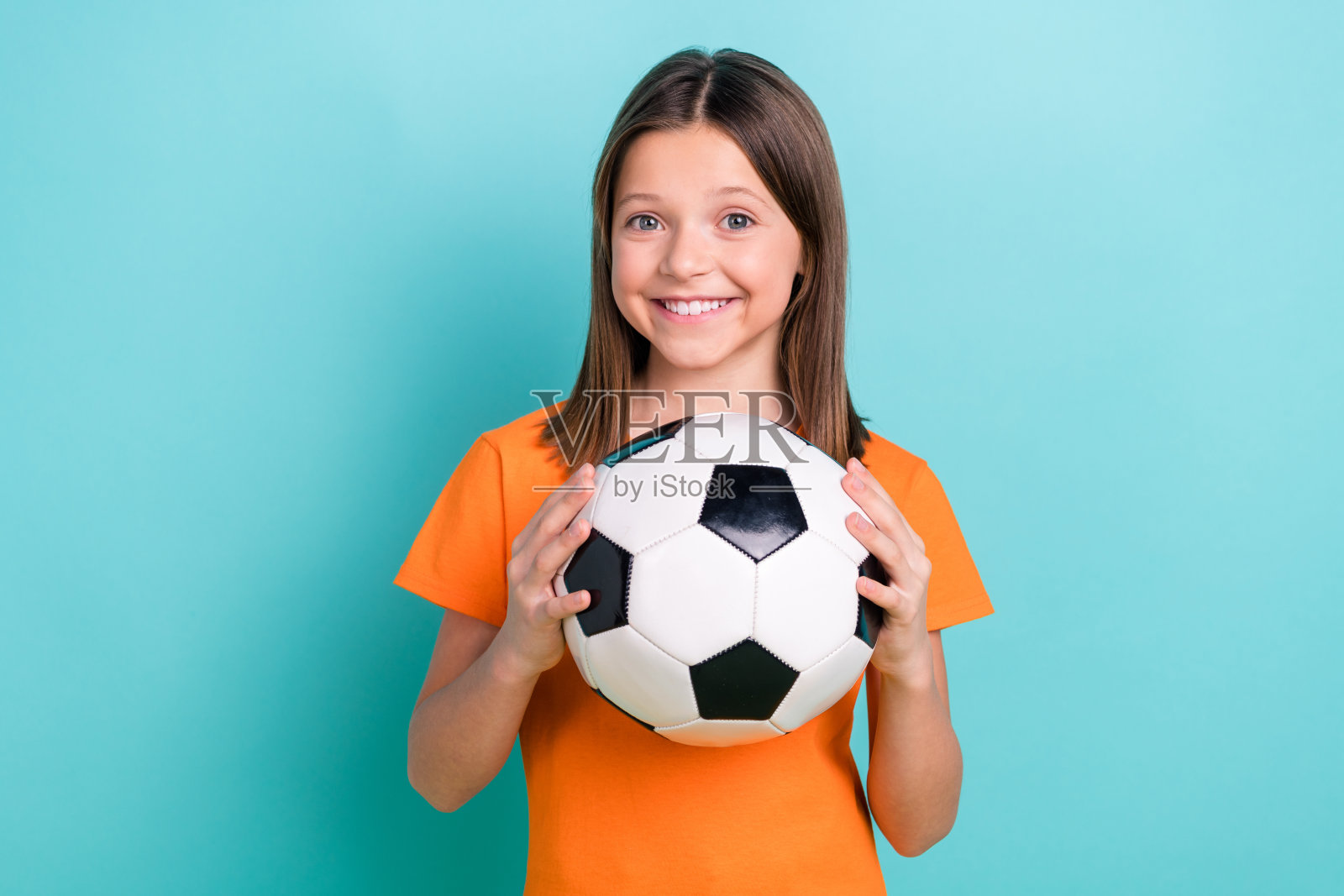 心情愉快的女学生肖像，直发，穿着橙色T恤，双手托着足球，背景为青绿色。照片摄影图片