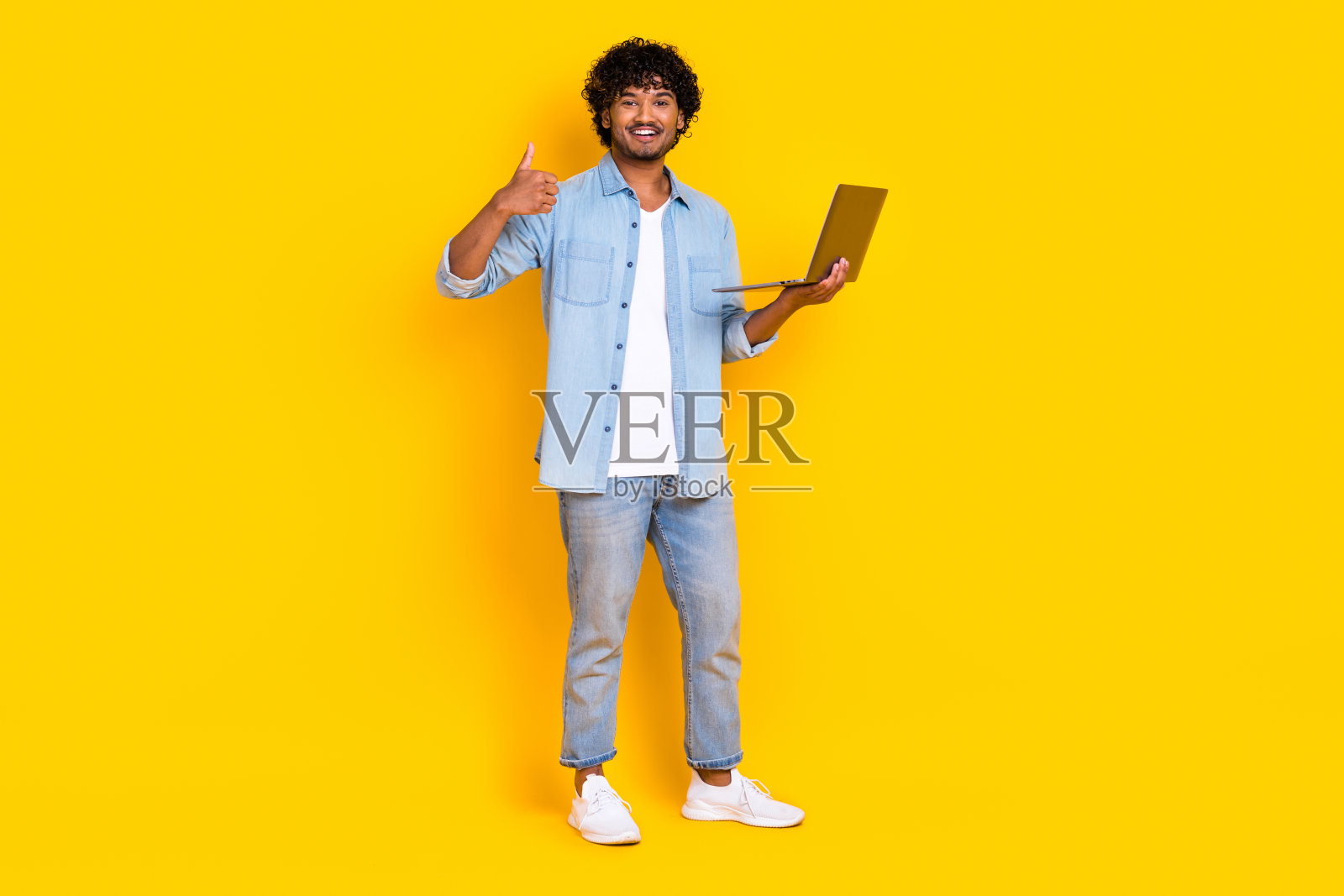 全身照，聪明的男人穿着牛仔衬衫和时尚的牛仔裤，手持笔记本电脑，竖起大拇指表示工作出色，背景为黄色。照片摄影图片