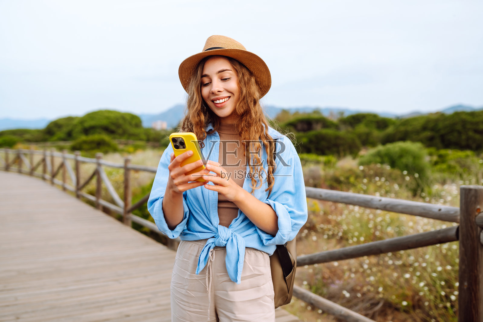一个可爱的游客戴着帽子，手里拿着手机，走在木栈道上照片摄影图片