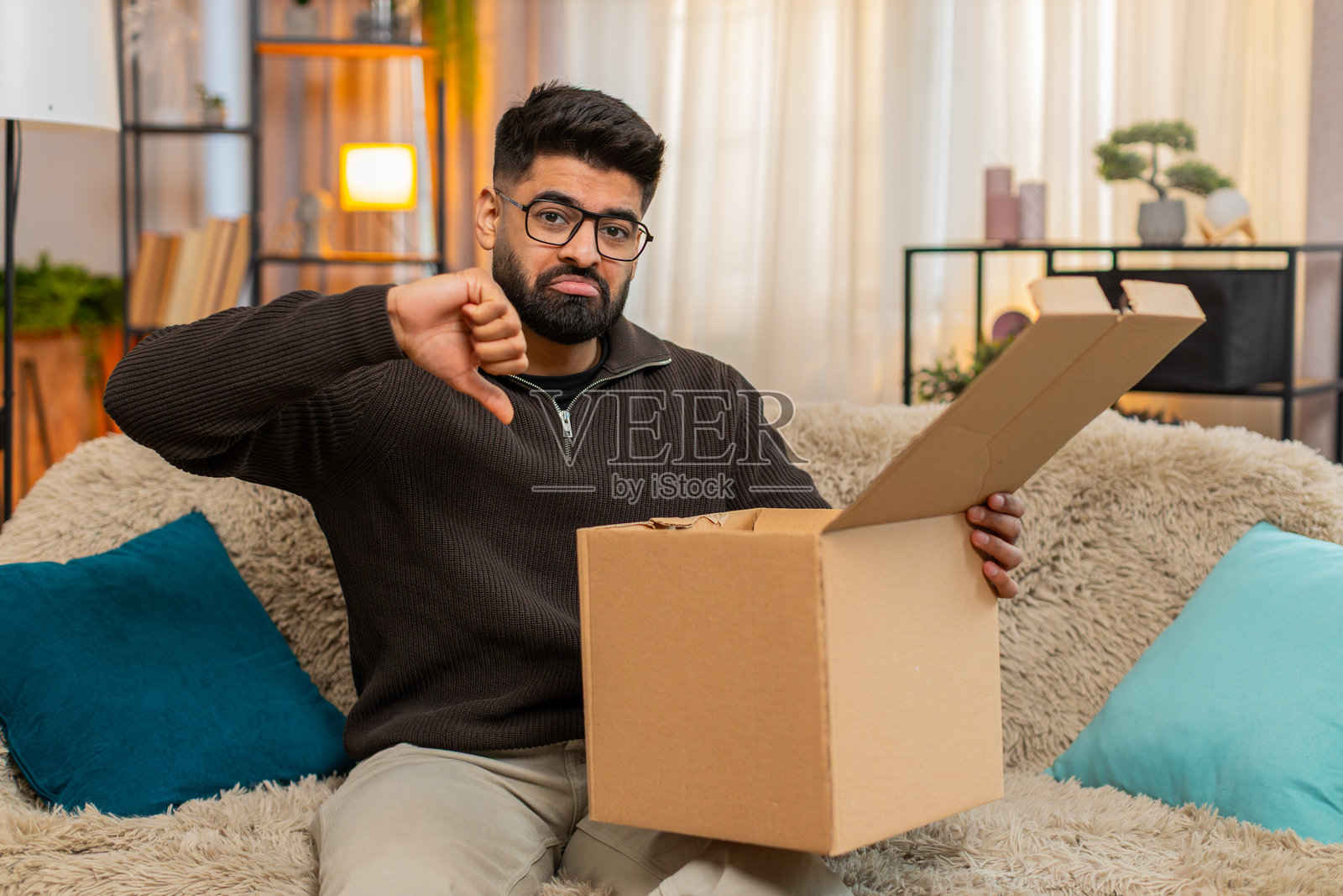 印度男子坐在沙发上打开盒子，发现网上购物订单中的物品错误，摇了摇头。照片摄影图片