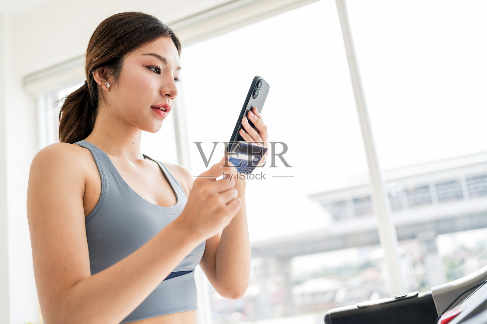 身材健美的年轻女性穿着运动装备，手持智能手机和信用卡，站在跑步机上，融合了健康与现代生活方式，象征着便利性、电子商务和健身动力。照片摄影图片