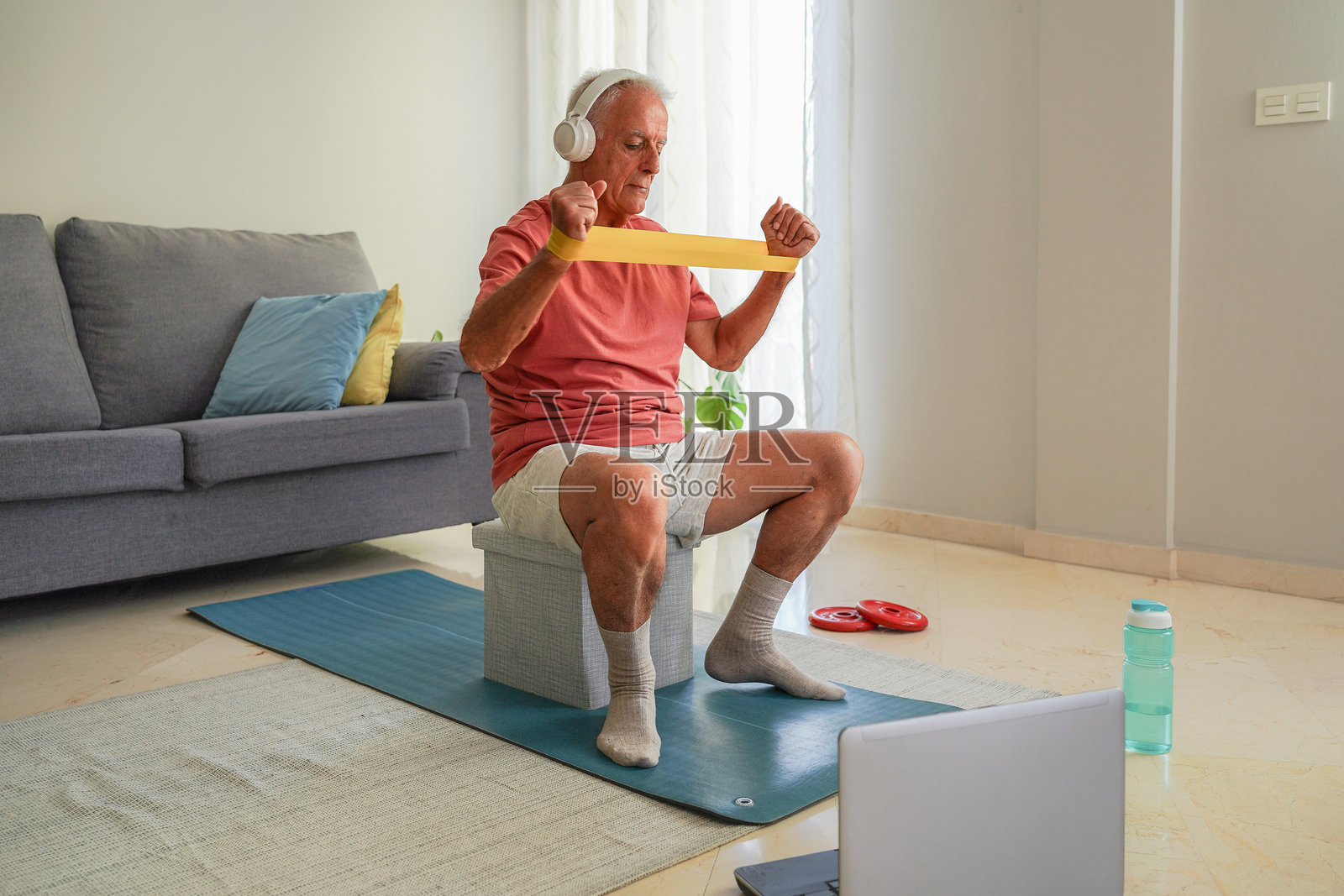 老年男子在家中戴着耳机，使用阻力带锻炼，跟随笔记本电脑上的在线健身课程。照片摄影图片