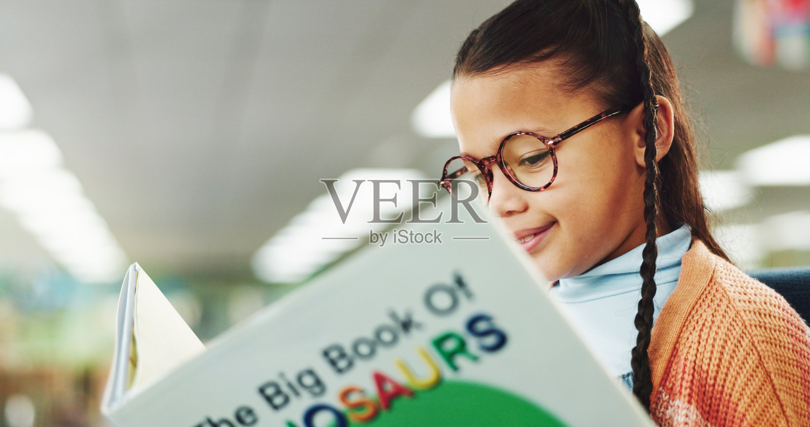 女孩在图书馆里微笑着阅读书籍，适合教育、学习或关于恐龙故事的眼镜。学校孩子，快乐并获得奖学金，适合语言、知识或学习以促进学术发展。照片摄影图片