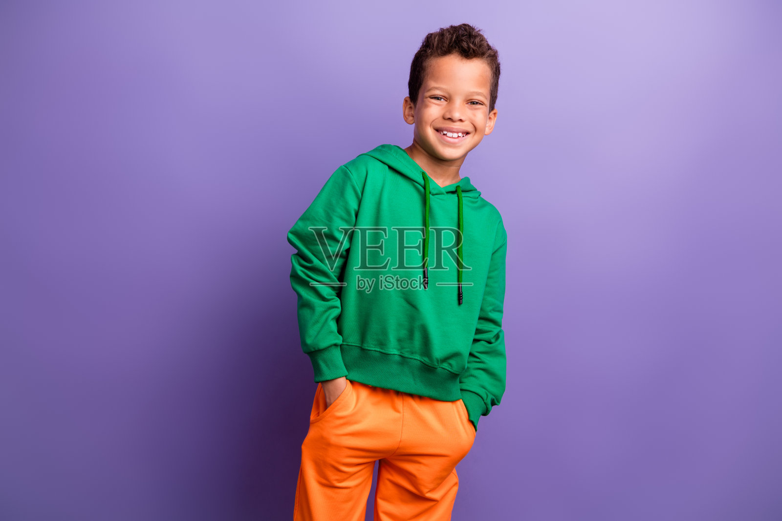 自信快乐的小男孩穿着绿色卫衣，微笑着走在紫色背景前。照片摄影图片