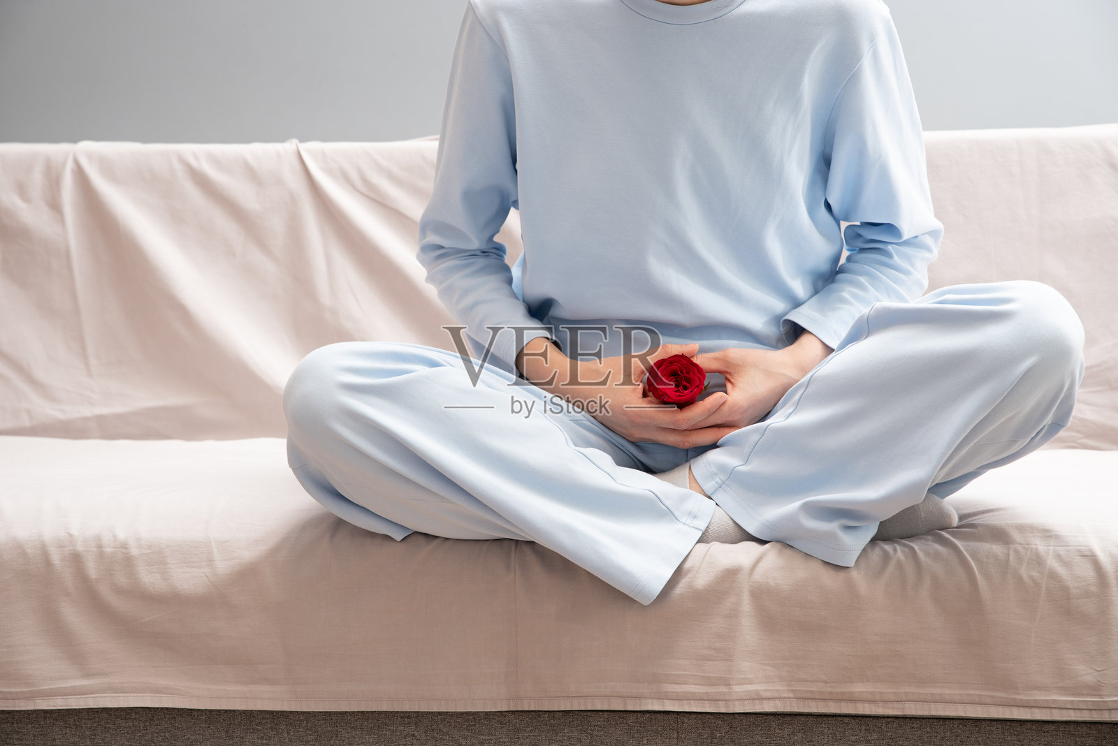 一位穿着蓝色睡衣的匿名女性坐在沙发上，手中握着一朵红色花蕾。月经期间腹痛。照片摄影图片