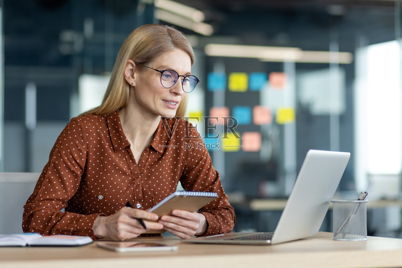一位戴眼镜的女商人在办公室桌前认真地使用笔记本电脑，旁边放着一本笔记本。照片摄影图片