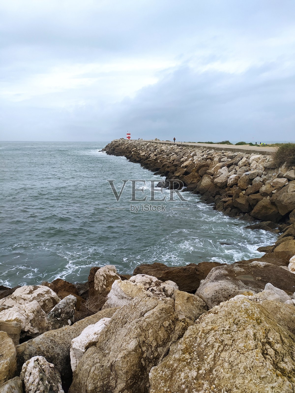 戏剧性的海岸场景，岩石防波堤延伸入海，乌云密布的天空下，远处的灯塔标志着地平线。非常适合旅行、天气或海景主题。照片摄影图片