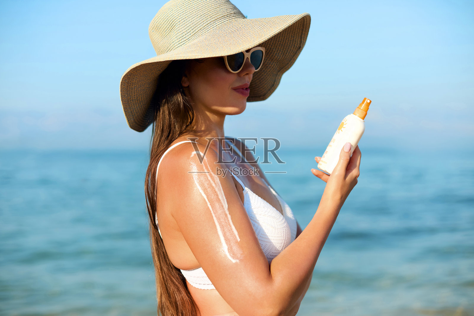 美丽的女性微笑着在皮肤上涂抹防晒霜。肌肤和身体护理概念。阳光保护。穿着比基尼的女孩在海滩上拿着防晒霜瓶。戴着帽子的女性涂抹保湿乳液。照片摄影图片