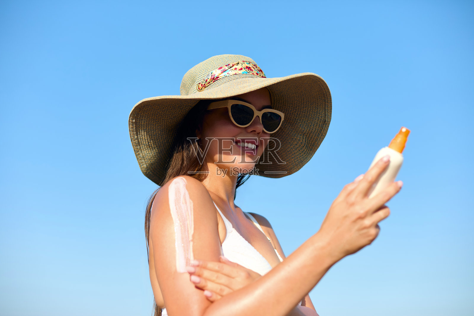 一位美丽的年轻女性在阳光明媚的海滩上享受着美好的一天，正在脸上涂抹防晒霜。她戴着太阳镜，头上戴着时尚的帽子，以保护肌肤免受夏日阳光的伤害。照片摄影图片