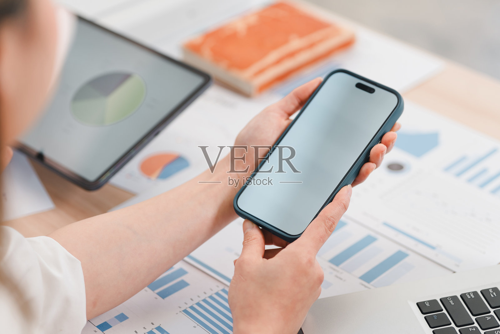 手持空白屏幕智能手机的人，周围是商业文件、图表和笔记本电脑，暗示工作或分析环境照片摄影图片