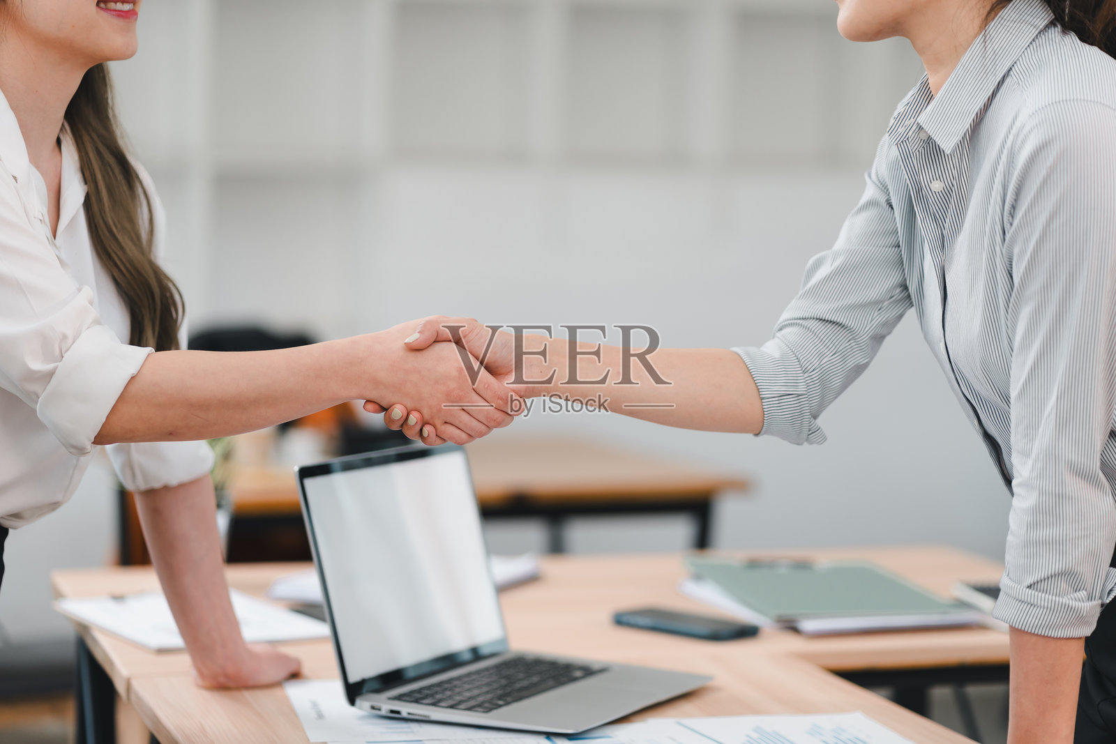 两位女商人在办公室环境中握手，象征着协议或合作，背景中有笔记本电脑和文件。照片摄影图片