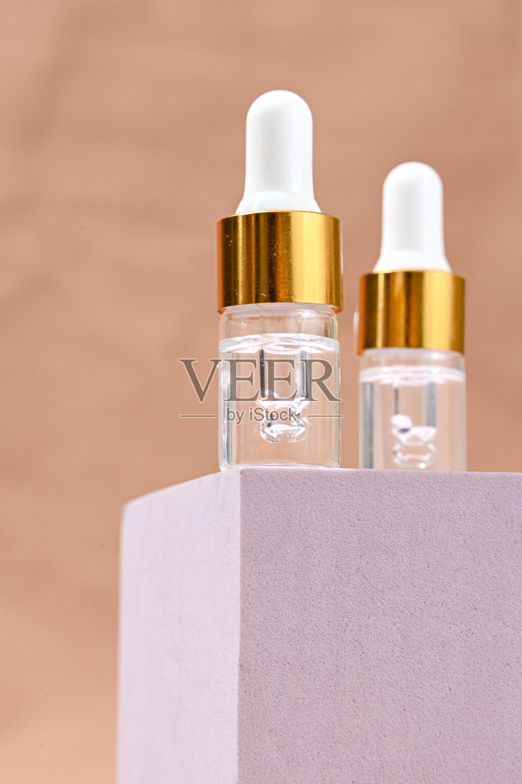两个透明玻璃精华滴管瓶，带有白色滴头和金色瓶颈，放置在柔和的粉色底座上，背景为桃色。极简护肤品牌，展现干净的美学。照片摄影图片