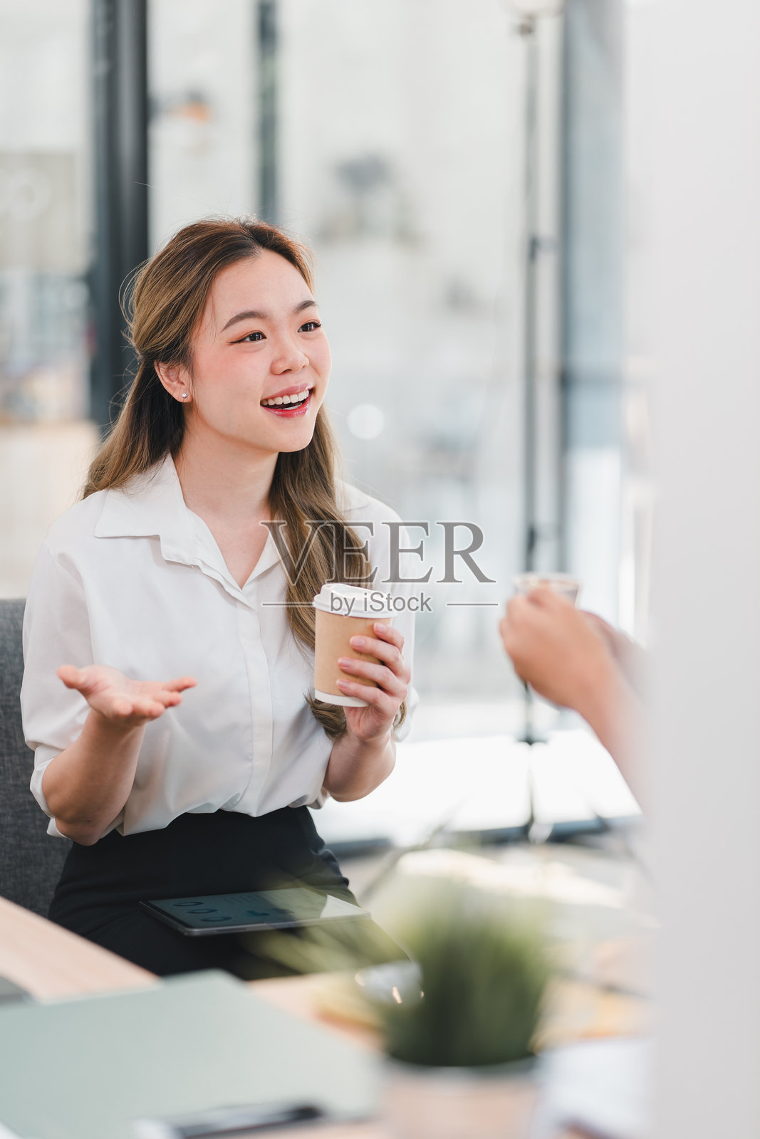 穿着白衬衫的快乐女性手持咖啡杯，在明亮的办公室环境中进行生动的对话，桌上有平板电脑，背景细节模糊。照片摄影图片